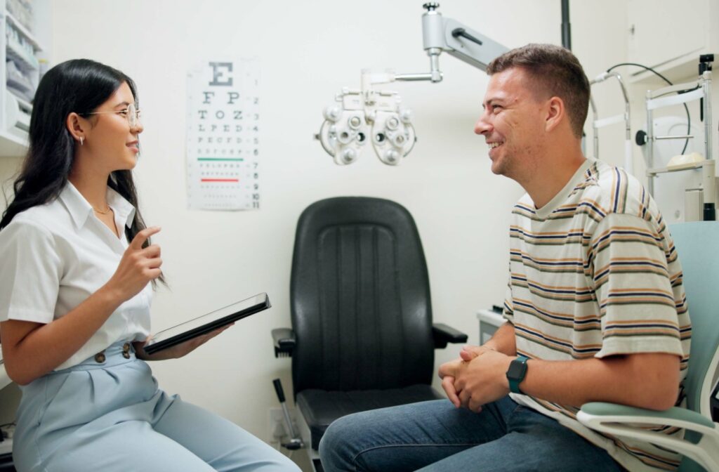 A patient sits in an exam room speaking with an optometrist holding a tablet, with an eye chart and exam equipment in the background.