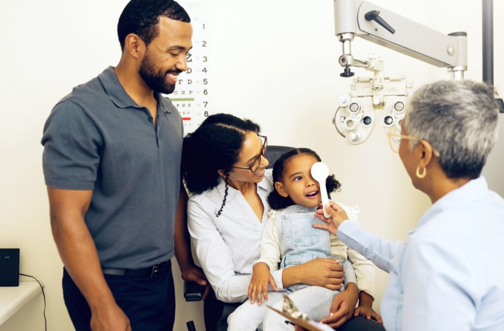 A child sits in their parents' lap while an eye doctor covers their left eye to assess their visual acuity.